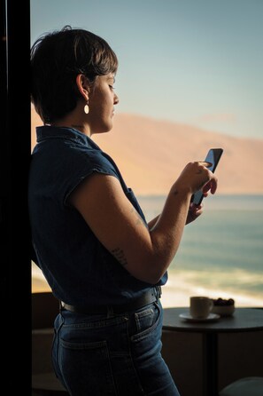 A person standing by a window with a view of the ocean, holding a smartphone.
