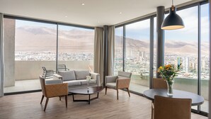 A modern hotel room with a large balcony, a sofa, two armchairs, a coffee table, and a dining table with a vase of flowers.