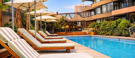 A swimming pool with lounge chairs and umbrellas, surrounded by greenery and a building with multiple floors.