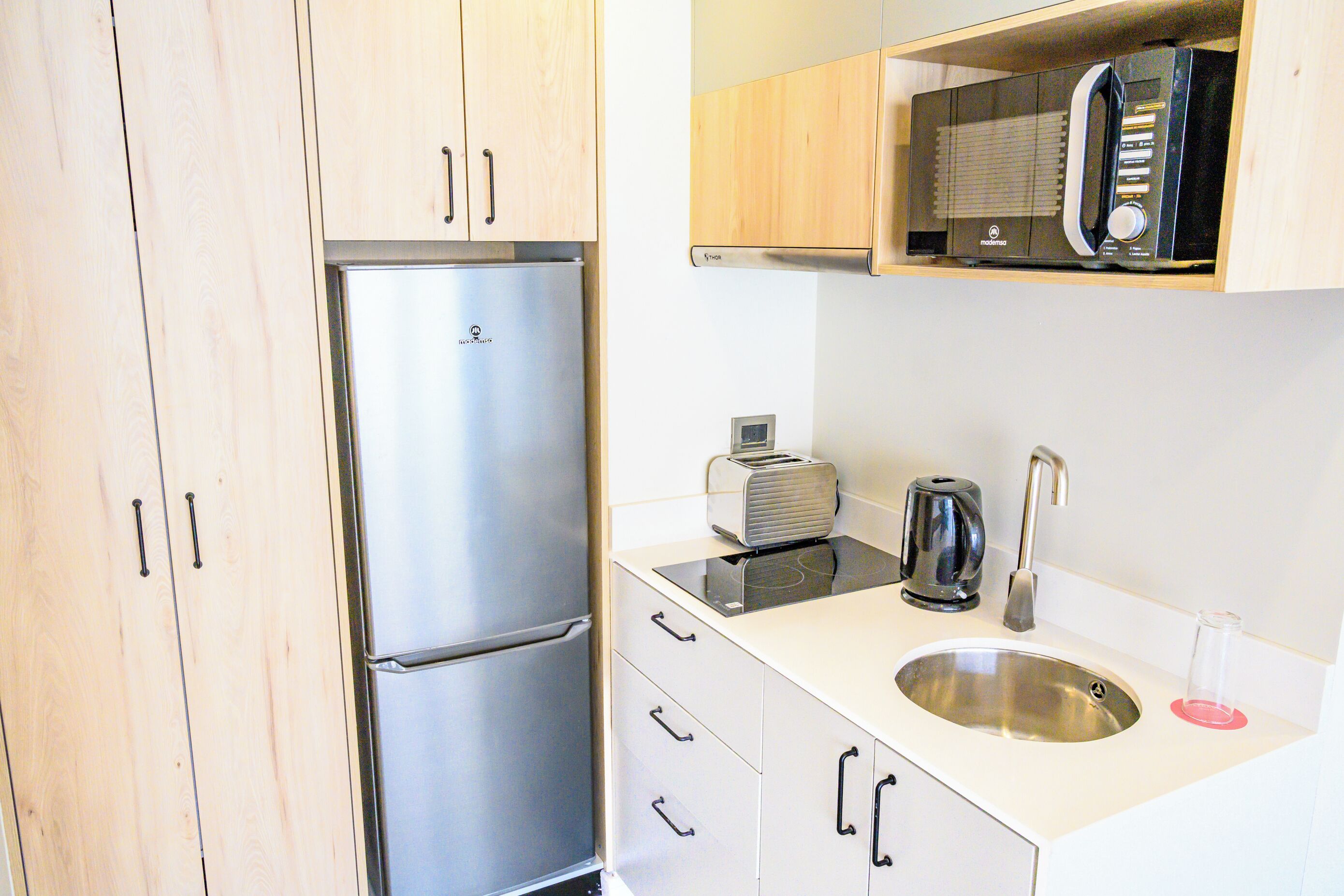A compact kitchen with a stainless steel refrigerator, a toaster, a kettle, and a sink.