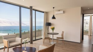 A modern hotel room with a large window overlooking the ocean, a round dining table, and a cozy seating area.