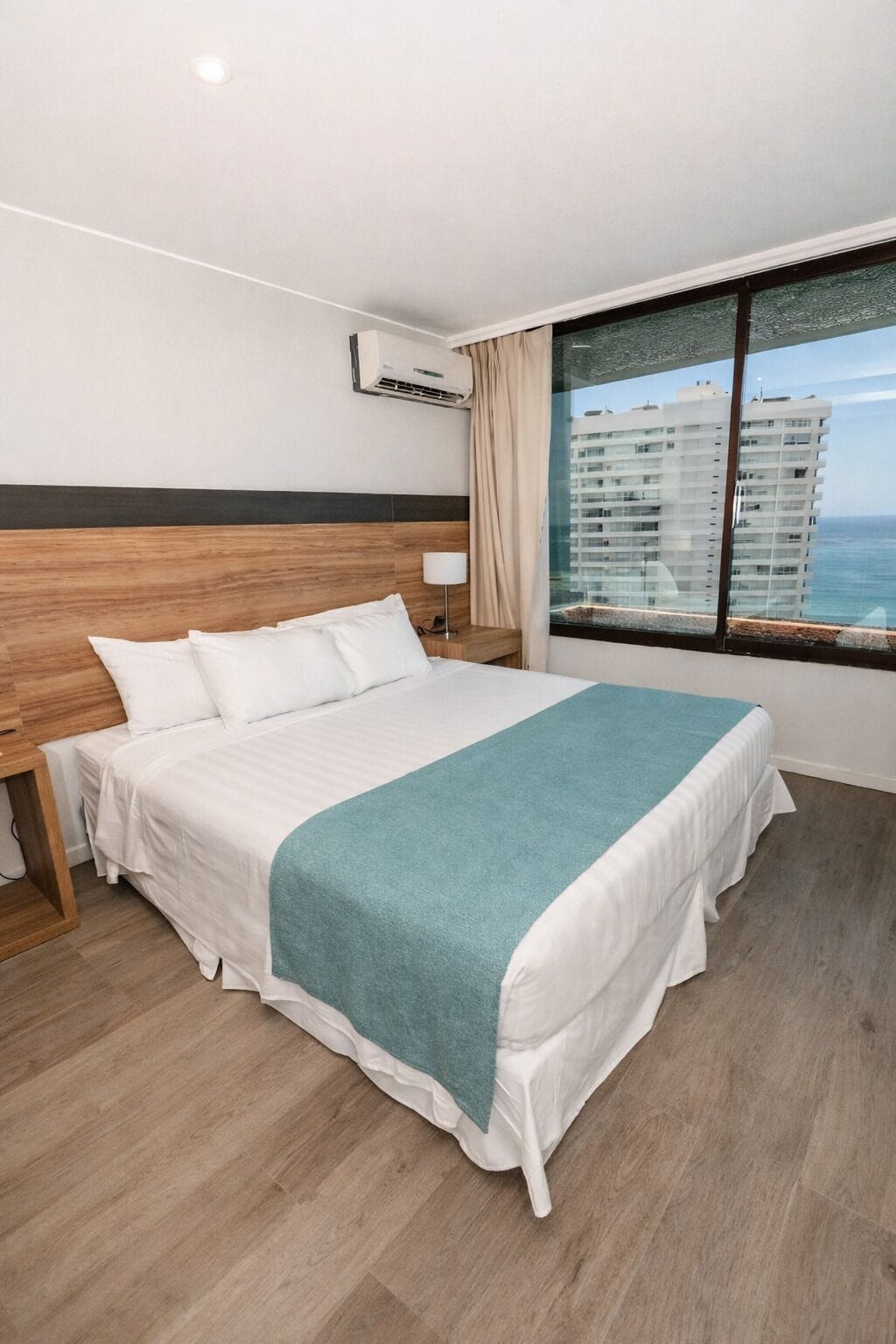 Une chambre d’hôtel avec un grand lit, une tête de lit en bois, une fenêtre donnant sur des immeubles et un climatiseur fixé au mur.