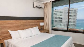 Une chambre d’hôtel avec un grand lit, une tête de lit en bois, une fenêtre donnant sur des immeubles et un climatiseur fixé au mur.