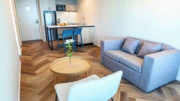 A modern living room with a grey sofa, a small round table with a vase, and a blue chair. A kitchen with a microwave and a sink is visible in the background.