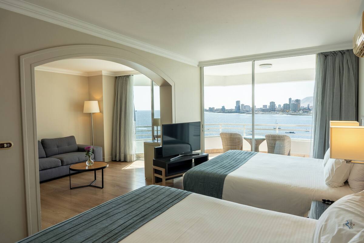 A hotel room with two beds, a TV, a sofa, a small table, and a view of the city and water.