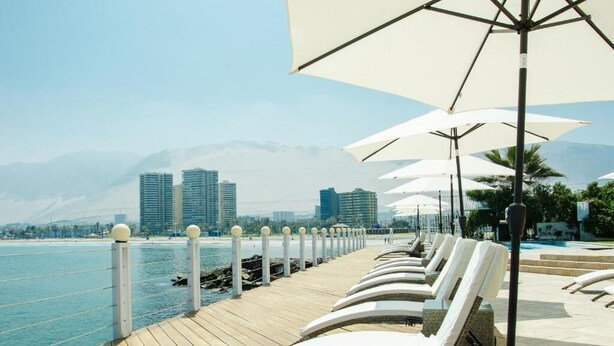 A waterfront promenade with sun loungers and umbrellas, a wooden deck, and a cityscape with mountains in the background. - Terrado Suites Iquique (Iquique)