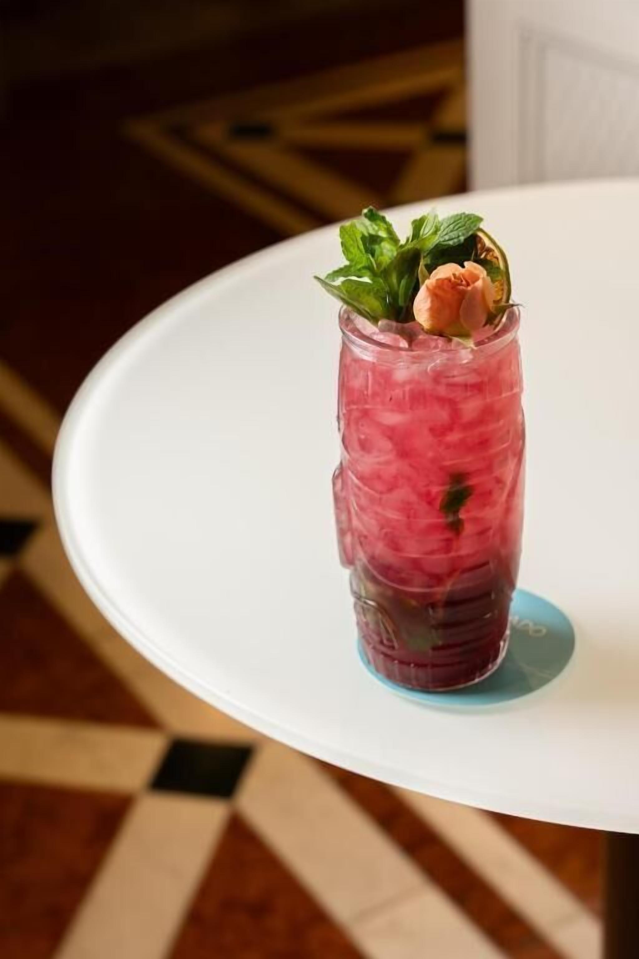 A glass with a pink beverage, garnished with mint and a peach slice, on a white table.