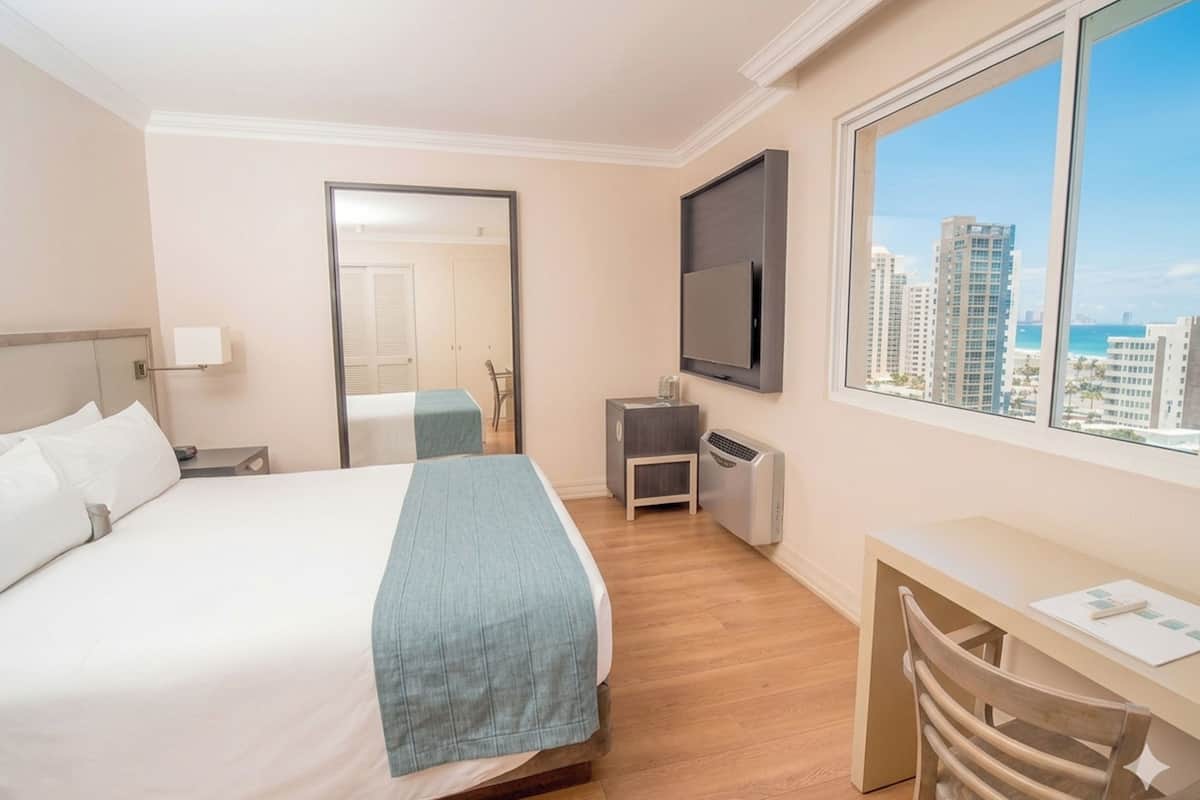 A hotel room with a bed, a desk, a chair, a TV, a window with a city view, and a mirror.