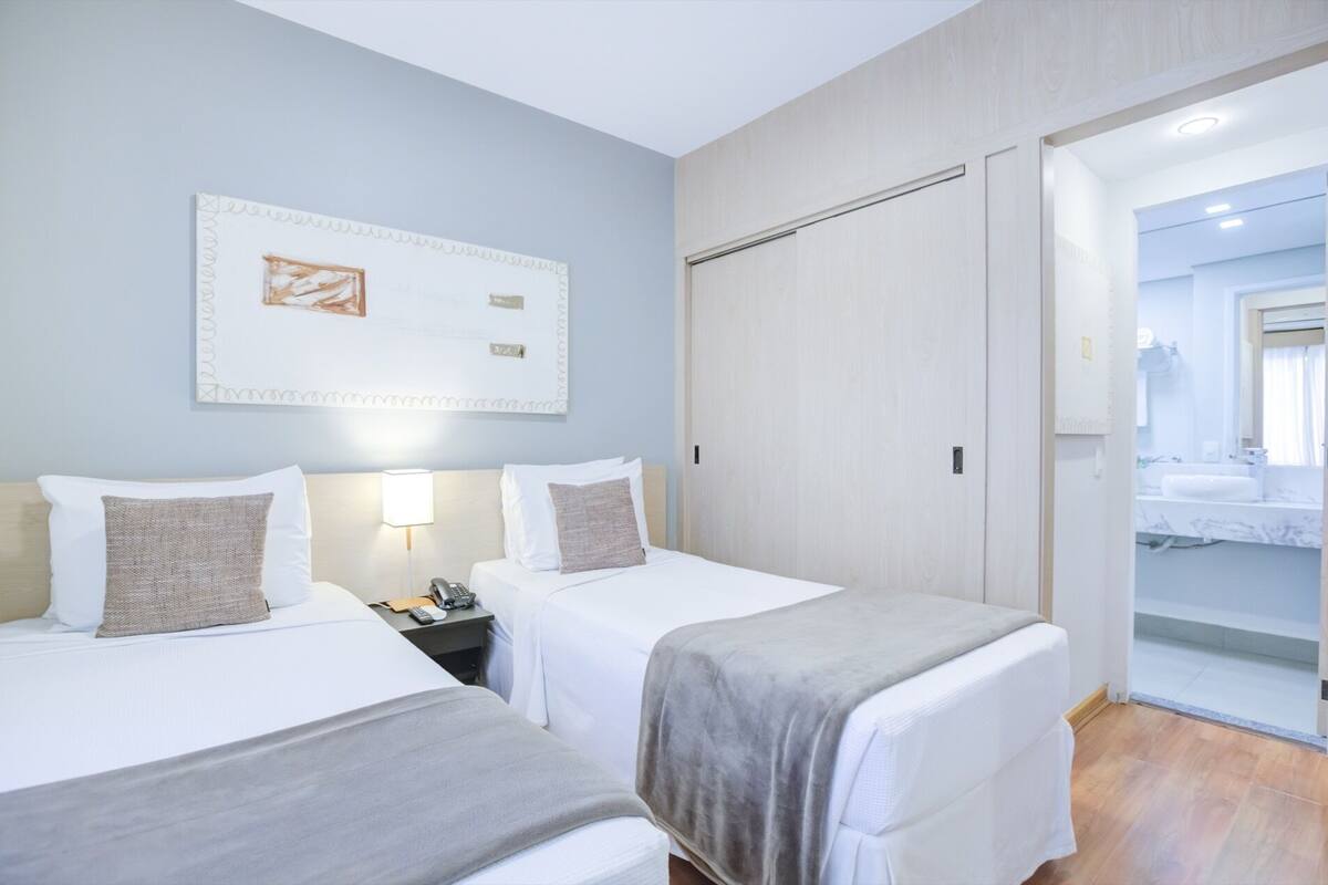A hotel room with two beds, a bedside table with a phone, a painting on the wall, and a bathroom visible through an open door.