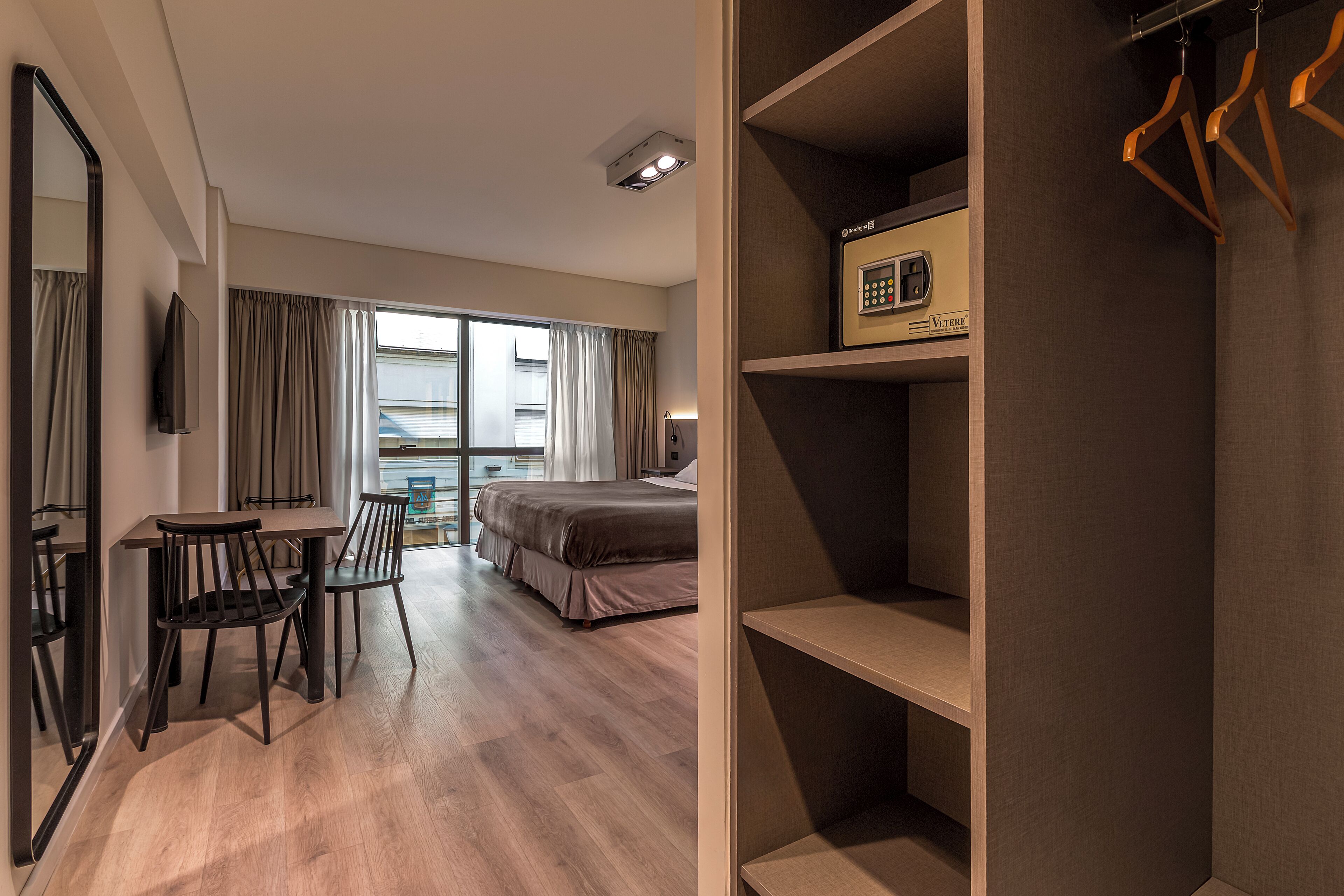 A hotel room with a bed, a desk with chairs, a wardrobe, and a window with curtains.