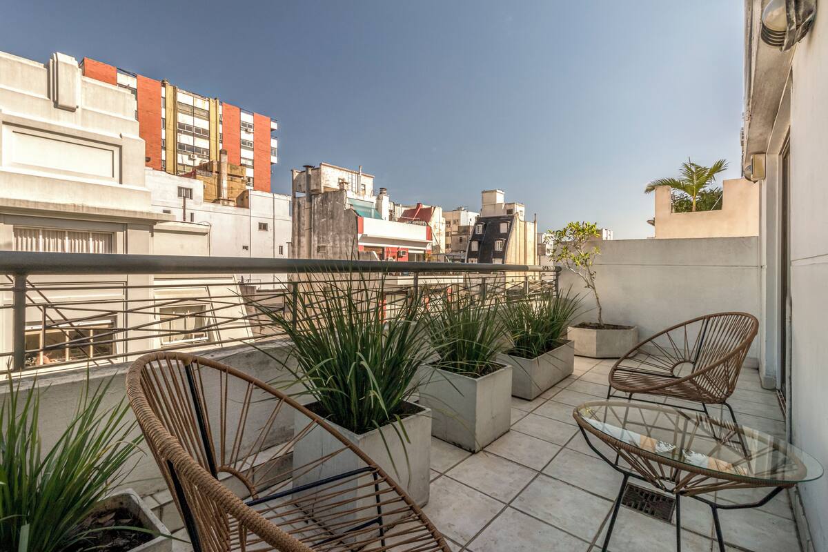 A rooftop terrace with wicker chairs, potted plants, and a glass table.