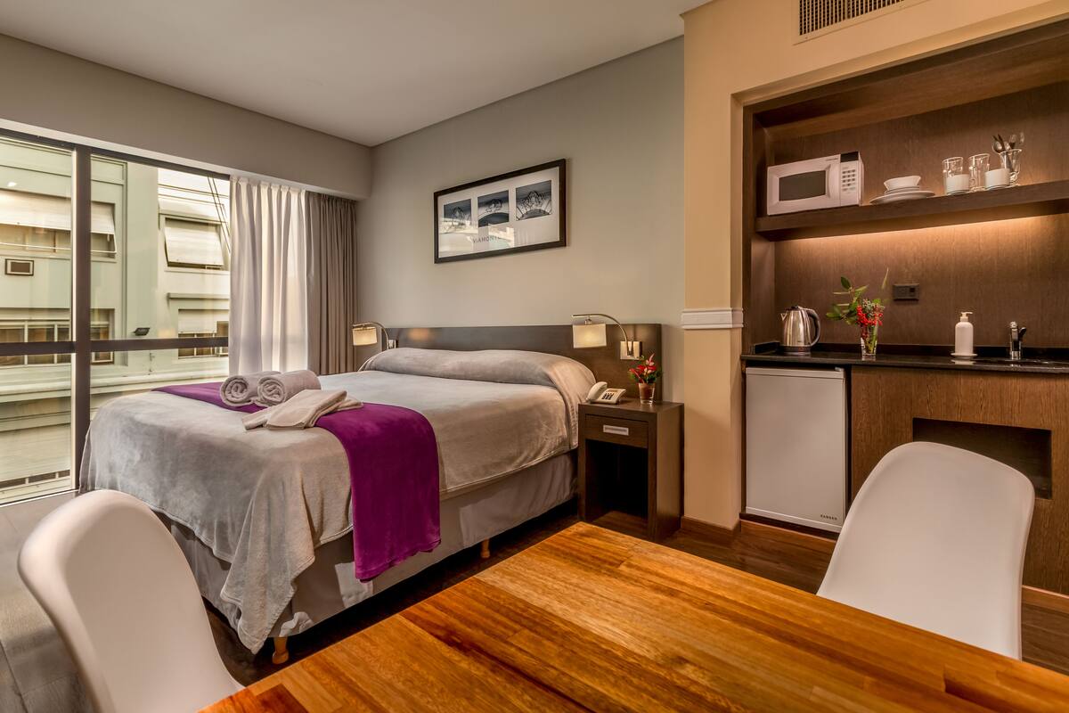 A hotel room with a bed, bedside tables, a desk, a chair, a microwave, a small refrigerator, and a window with curtains.