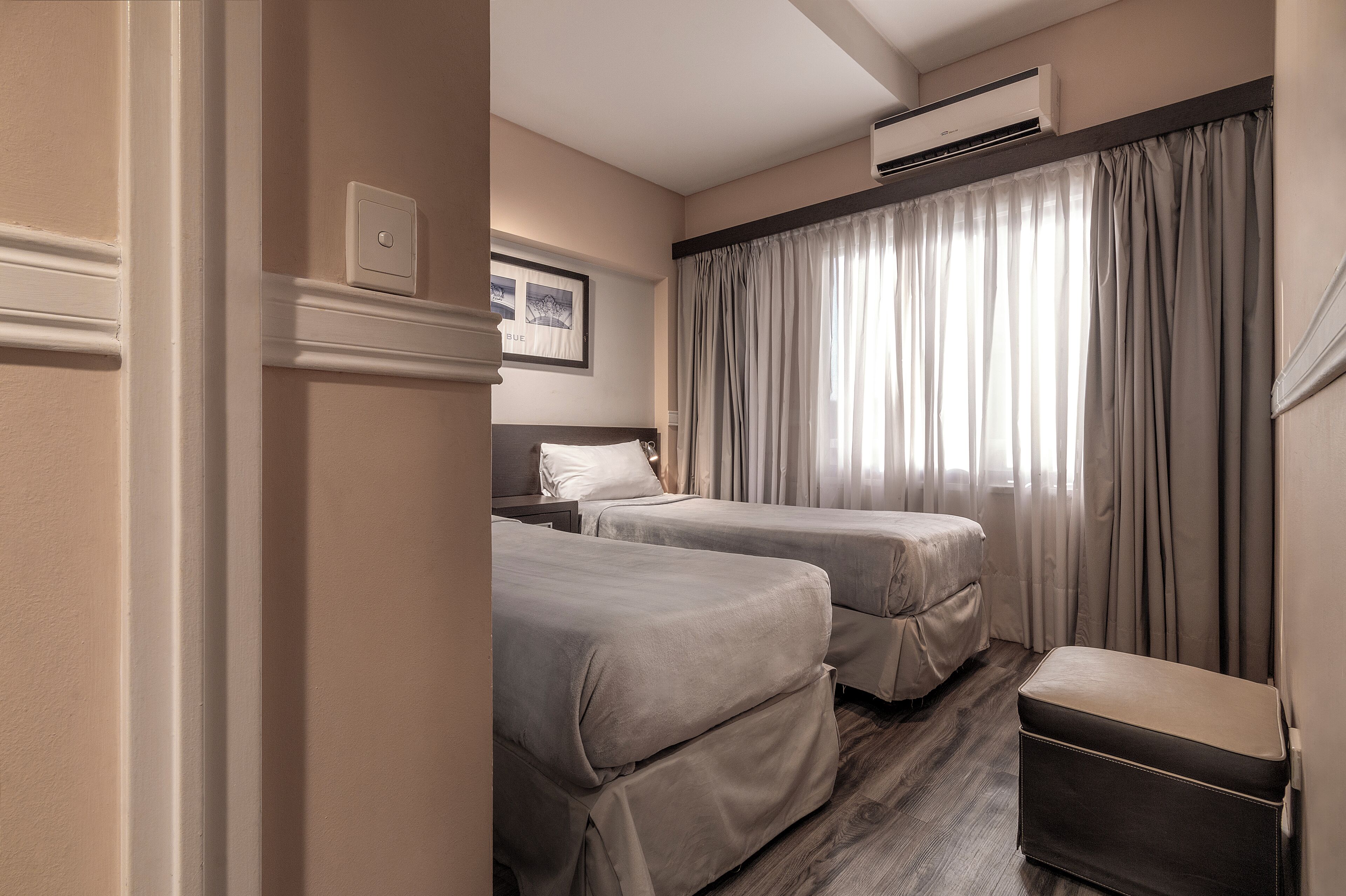 A hotel room with two beds, a bench, an air conditioning unit, and a window with curtains.