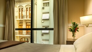 A hotel room with a large bed, a view of a building with ornate balconies, a vase with flowers on a nightstand, and a lamp. - Up Viamonte Hotel (Buenos Aires)