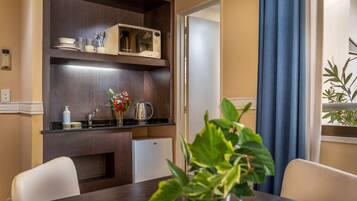 Una cocina funcional con microondas, cafetera y un refrigerador pequeño. Se aprecia una mesa de comedor con sillas y una planta en maceta al frente.