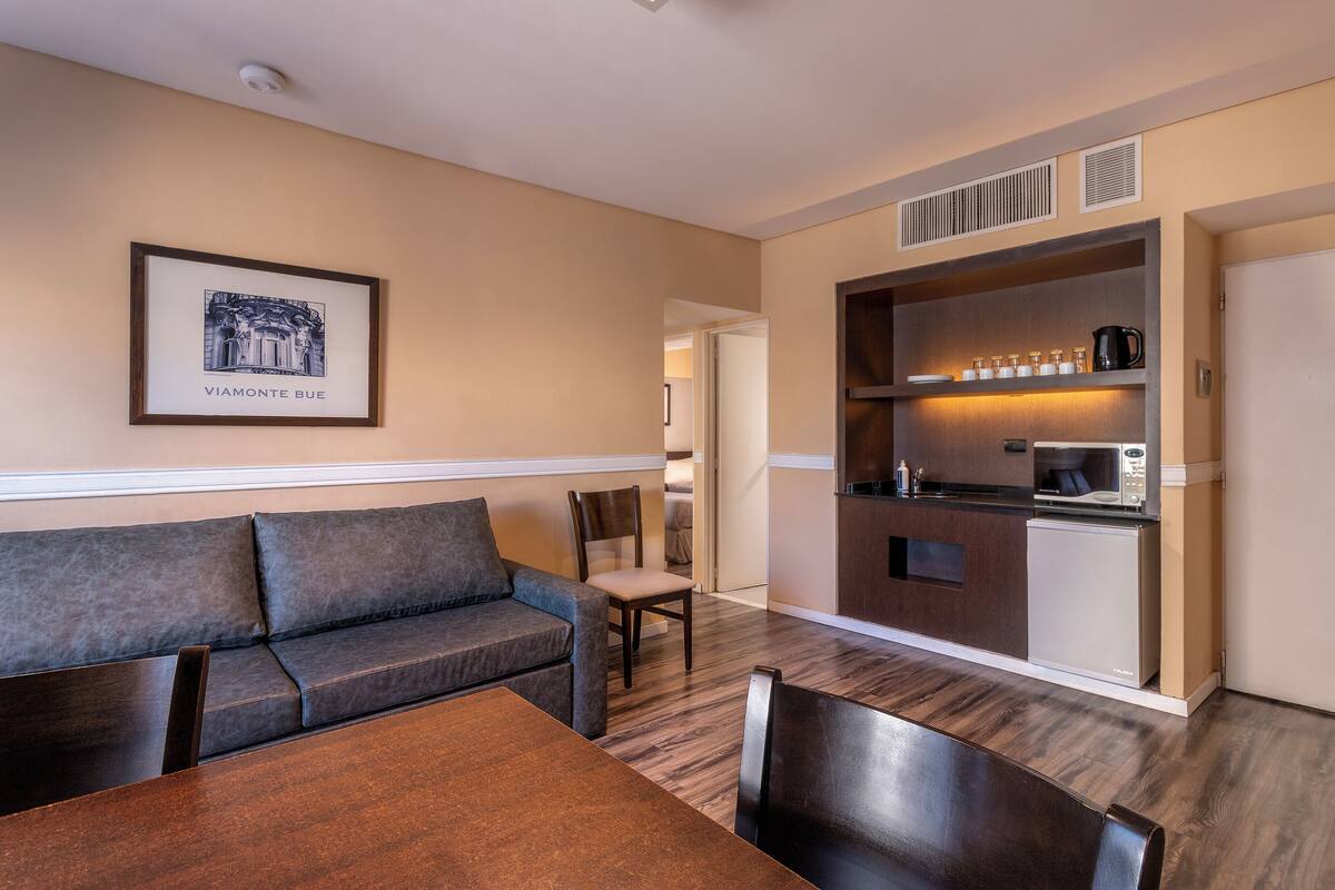 A modern hotel room with a sofa, dining table, and kitchenette.