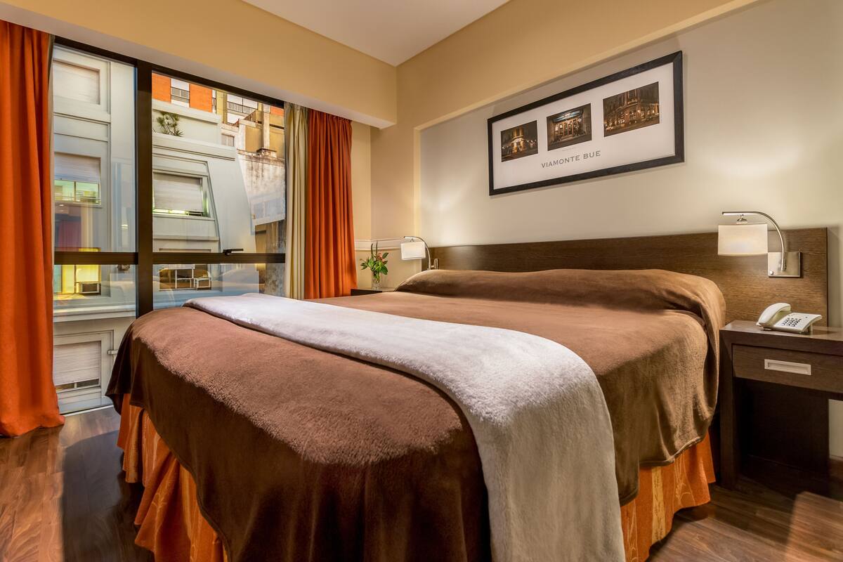 A hotel room with a large bed, a nightstand with a lamp, a framed picture on the wall, and a window with curtains.