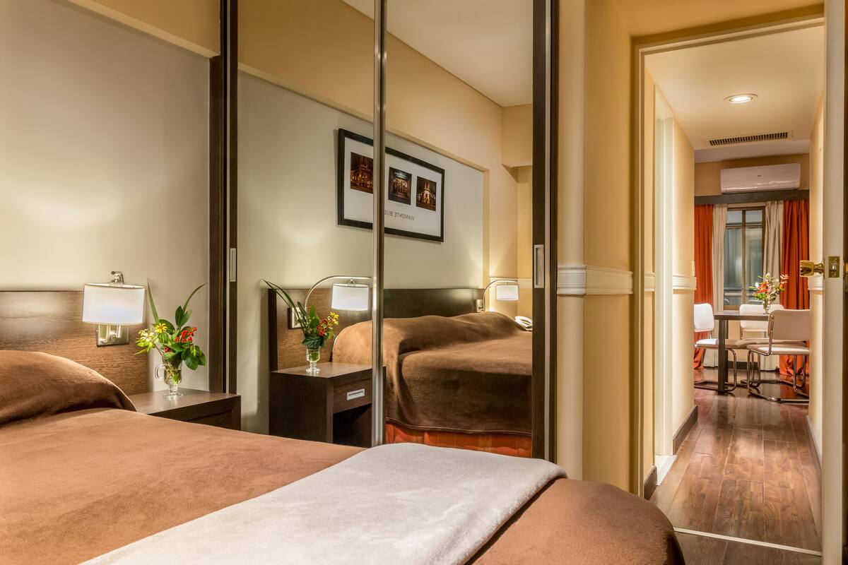 A hotel room with a large bed, bedside tables with lamps, a mirror, and a view of the dining area.