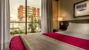 Een hotelkamer met een groot bed, nachtkastje, lamp en een raam met gordijnen.