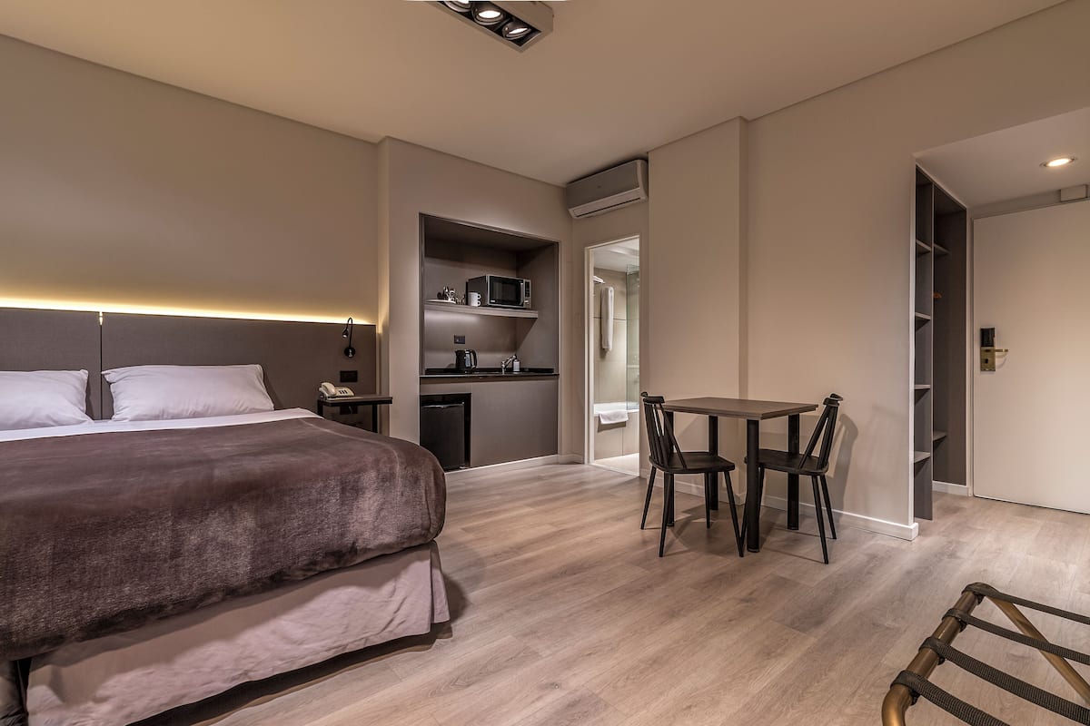 A modern hotel room with a large bed, a small kitchenette, a dining area with a table and chairs, and a separate bathroom.