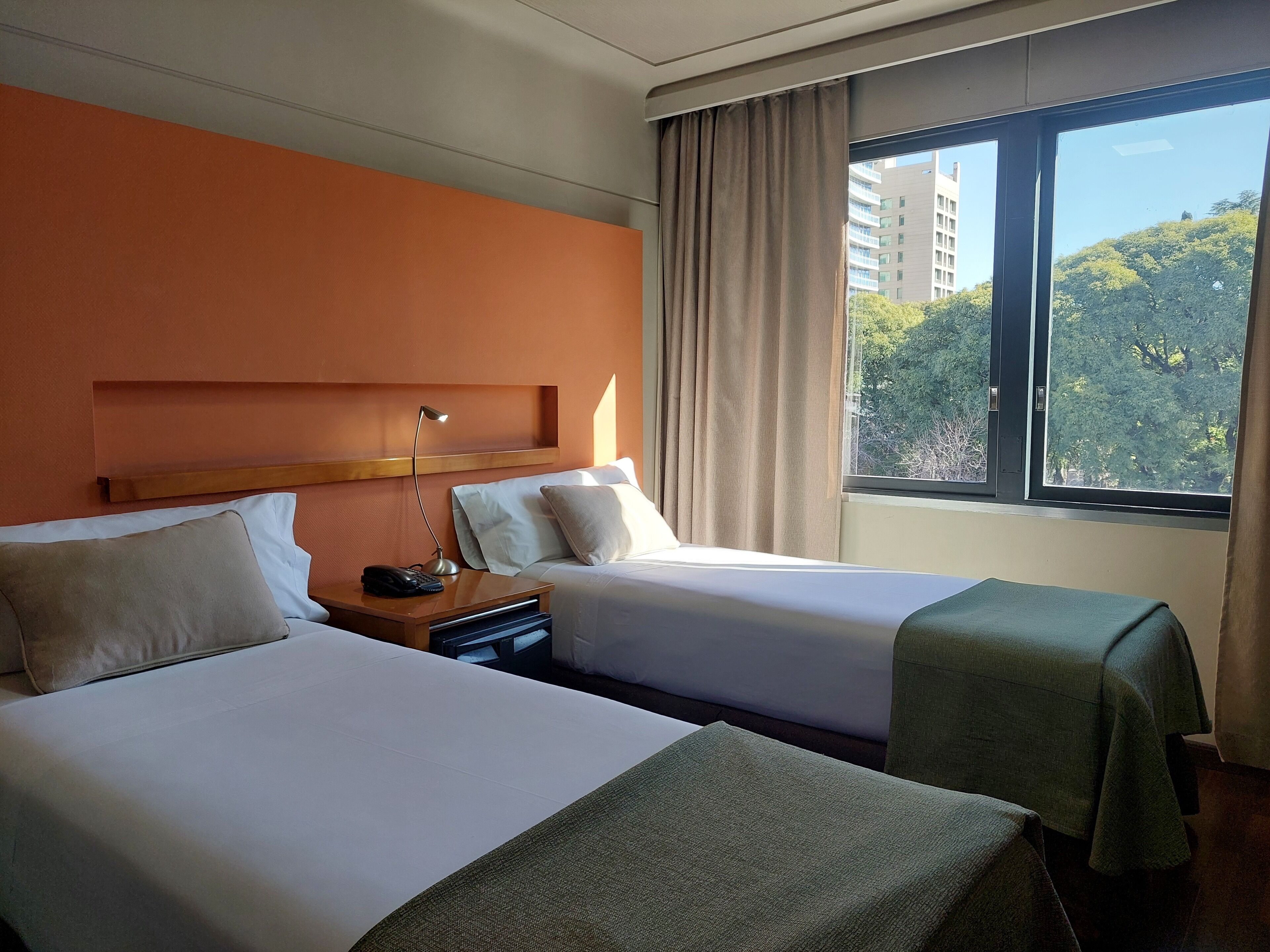 Habitación de hotel con dos camas, una mesita de noche con lámpara y ventana con vistas a árboles y edificios.