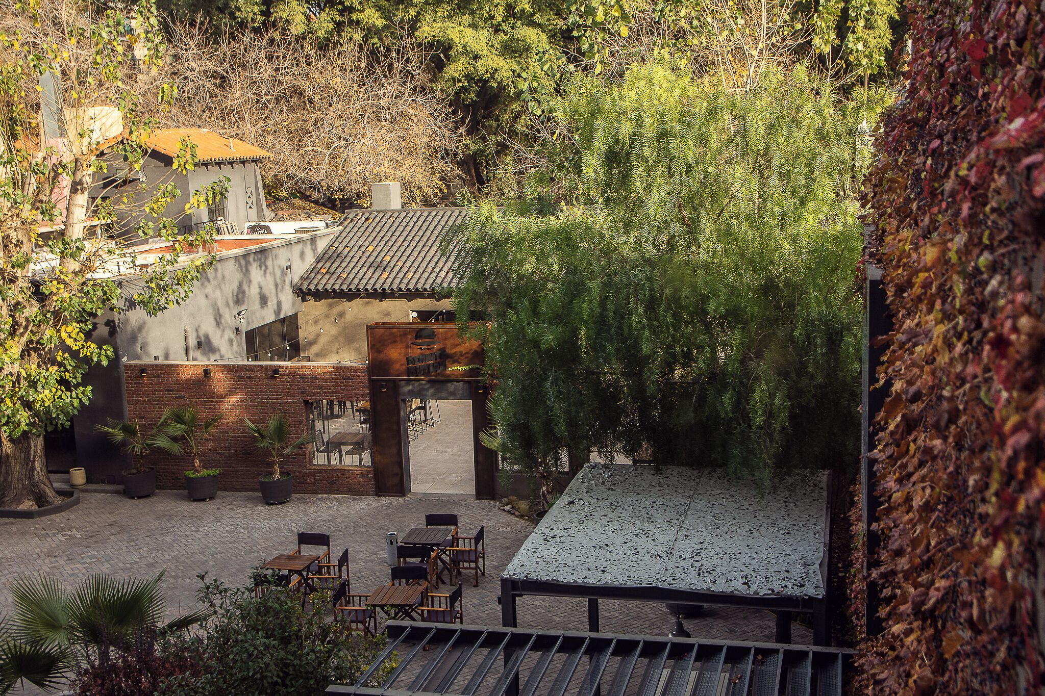 An outdoor seating area with tables and chairs, surrounded by trees and a building with a tiled roof.