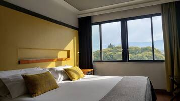 A hotel room with a large bed, a headboard, a window with a view of trees and mountains, and a curtain.
