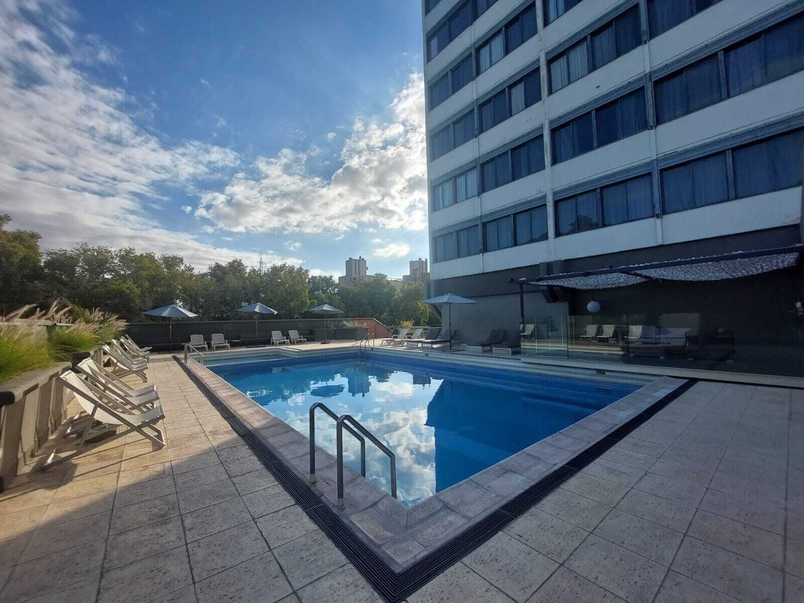 Piscina con sillas de descanso y sombrillas, rodeada por un edificio con múltiples ventanales.