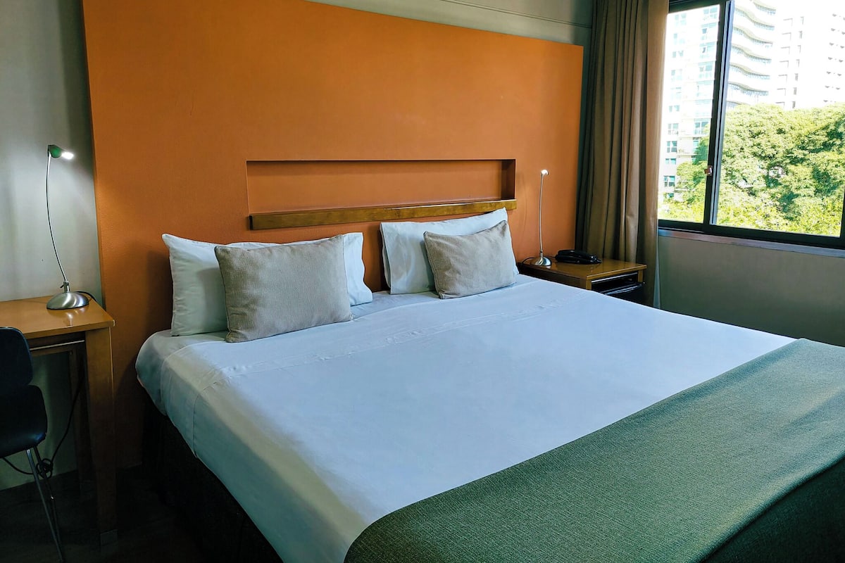 A hotel room with a large bed, a desk with a lamp, and a window with a view of buildings and trees.