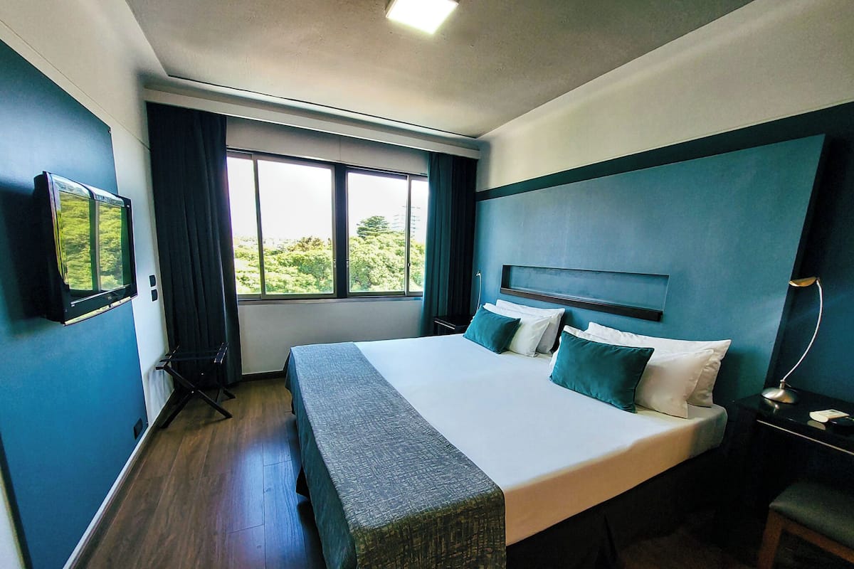 A hotel room with a large bed, a bedside table, a chair, a window with a view of greenery, and a wall-mounted TV.