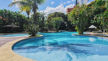 Une piscine dont les bords sont recouverts de carreaux, entourée de palmiers et de verdure, avec un bâtiment moderne en arrière-plan.