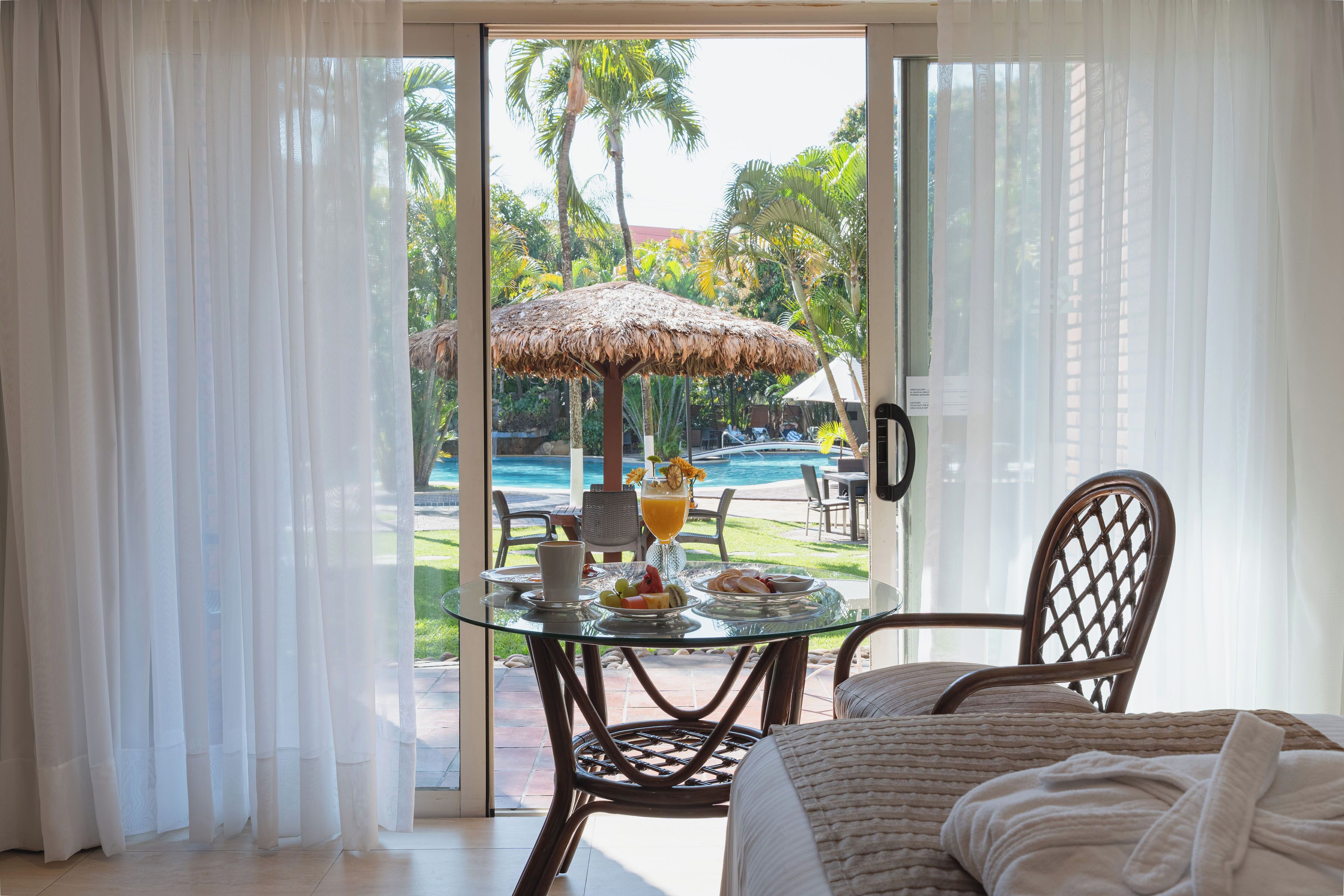 A room with a glass table and chairs, a bed, and a view of a pool and tropical vegetation.