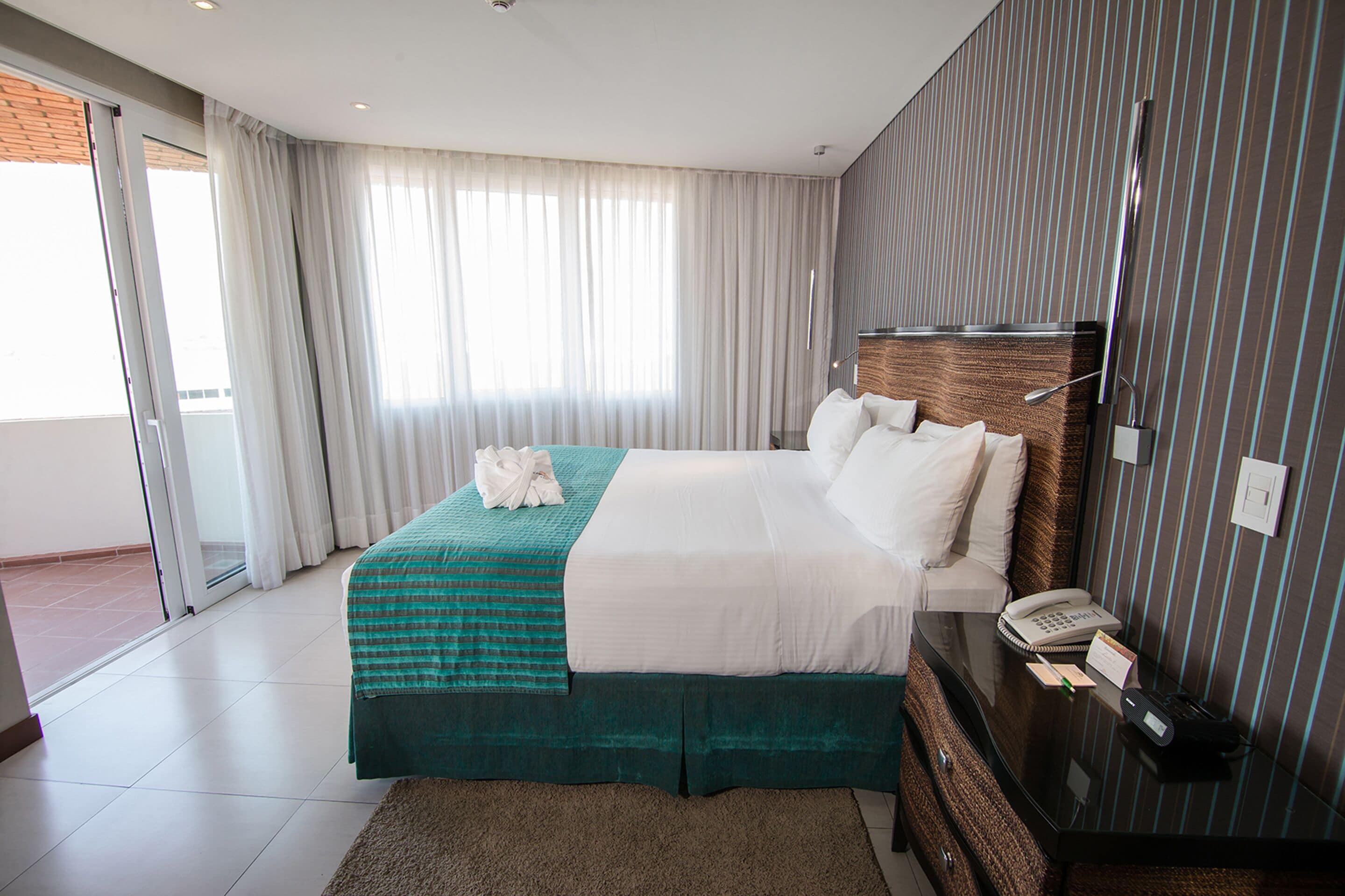A hotel room with a bed, a bedside table with a phone and a notepad, a window with curtains, and a view of the outside.