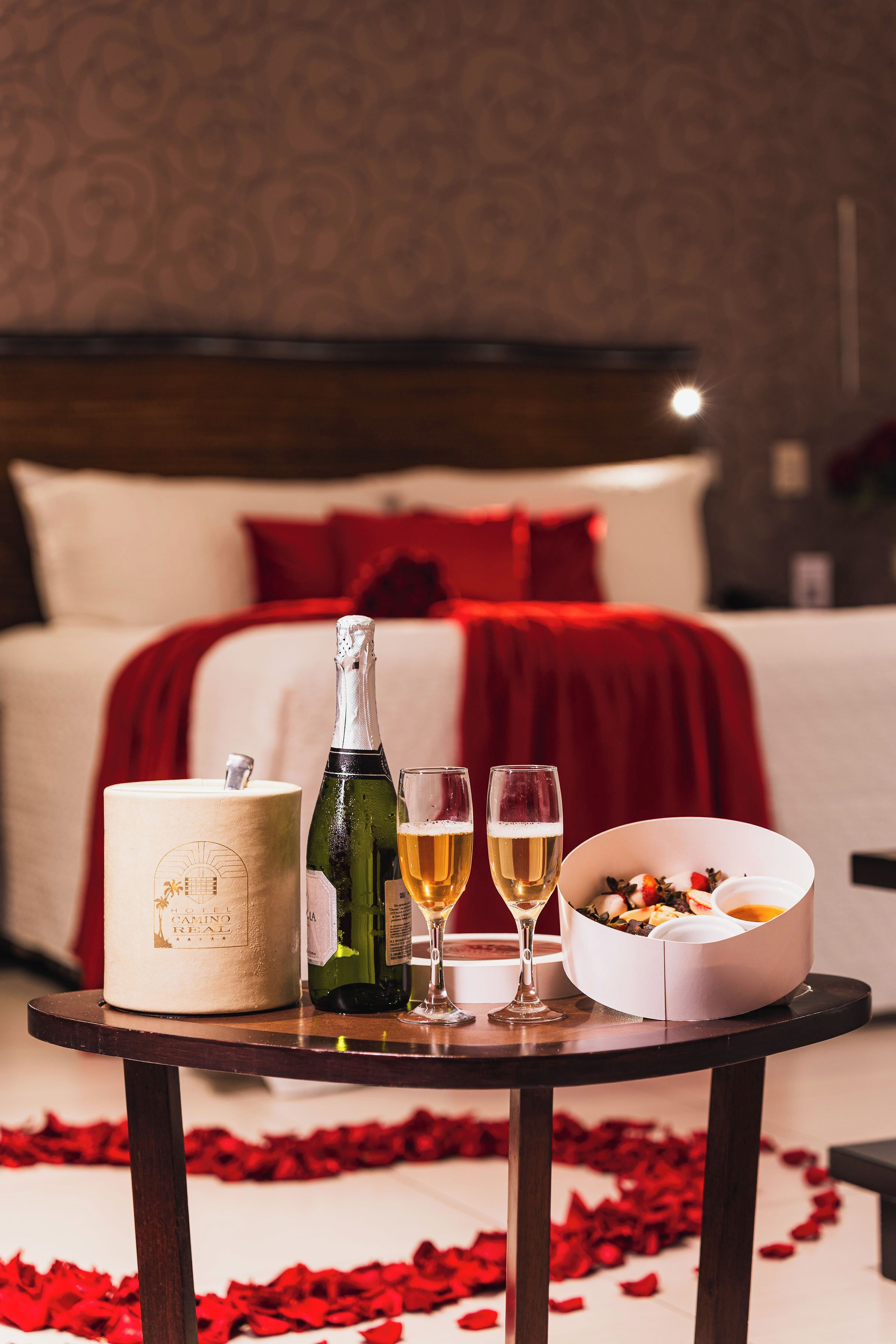 A hotel room with a bed, a table with a bottle, glasses, and a bowl of fruit.