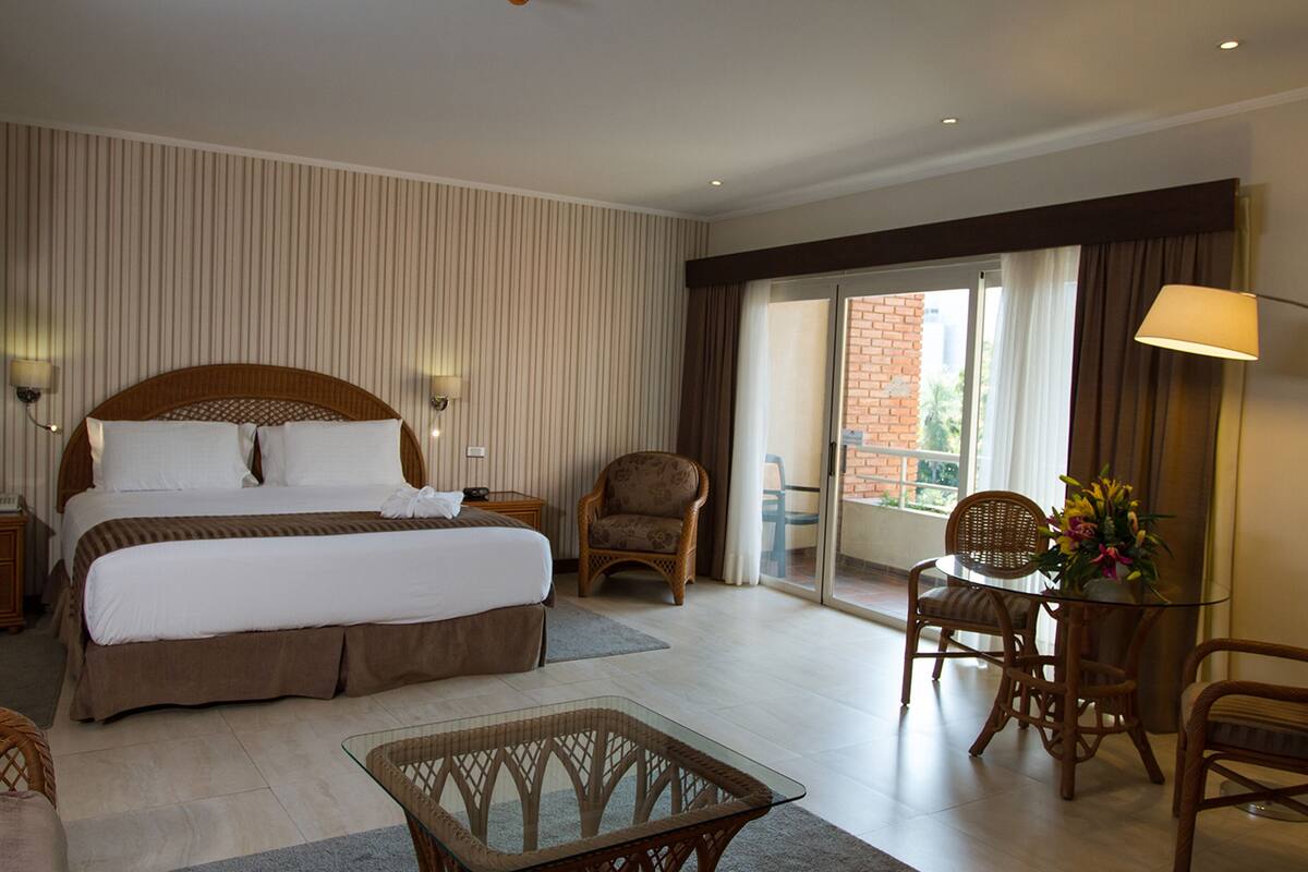 A hotel room with a large bed, a glass coffee table, wicker chairs, and a view of the outdoors.