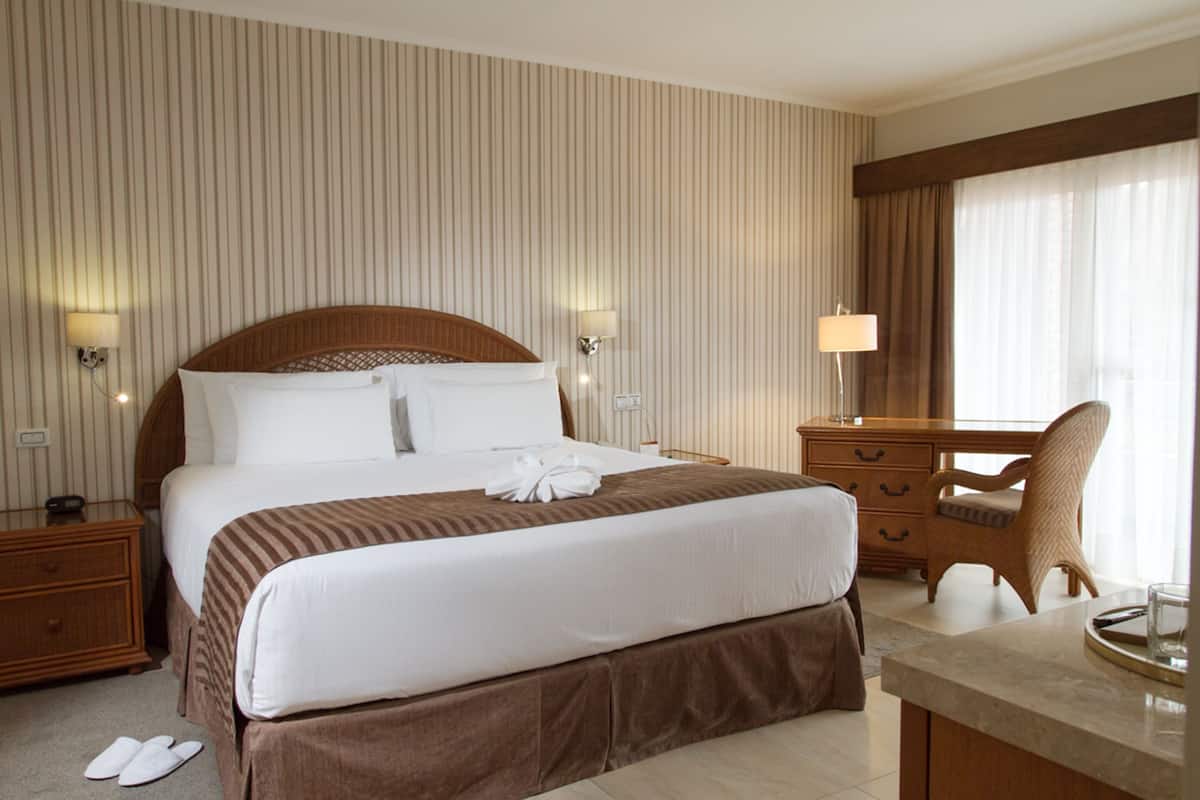 A hotel room with a large bed, a wooden nightstand, a desk with a chair, and a window with curtains.