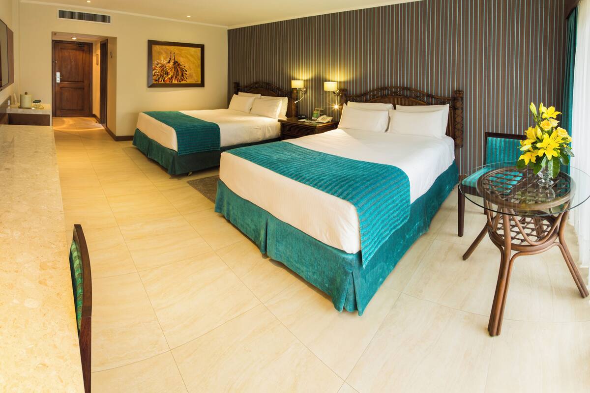 A hotel room with two beds, a sitting area with a glass table, and a view of the hallway.