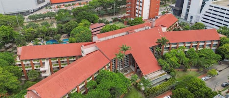 A multi-story building with a red roof, surrounded by greenery and adjacent to a commercial area.