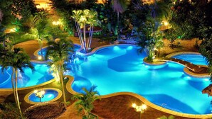 Une grande piscine illuminée, entourée de palmiers et bordée de sentiers.