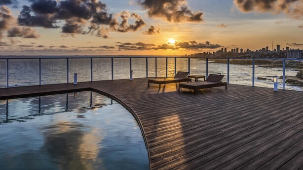 A wooden deck with lounge chairs and a pool, overlooking a city skyline at sunset. - Novotel Salvador Rio Vermelho (Salvador)