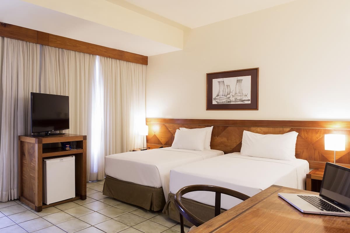 A hotel room with two beds, a TV, a small fridge, a desk with a laptop, and a framed picture on the wall.
