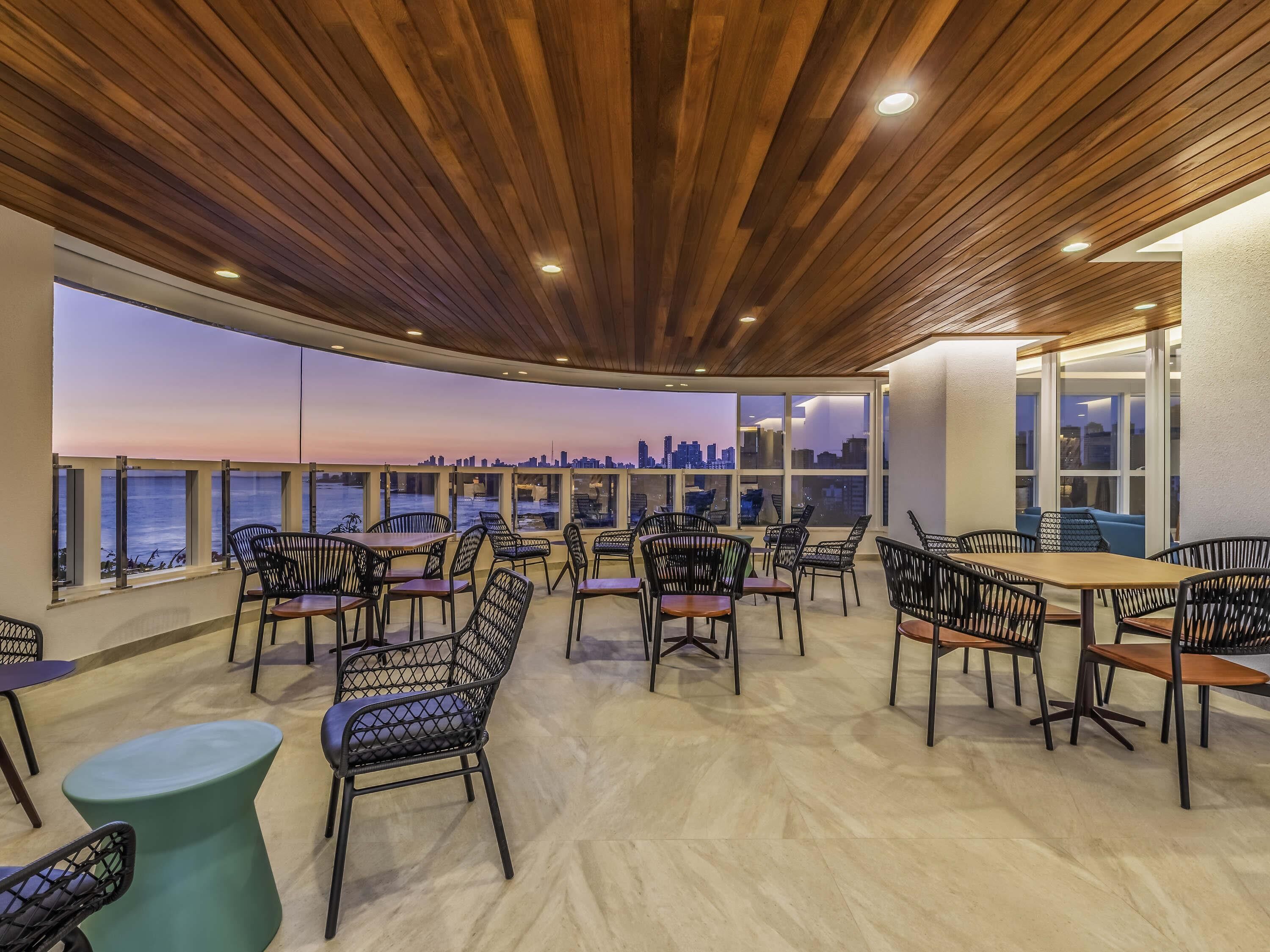 A rooftop terrace with wooden ceiling, seating, and a view of the city skyline at dusk.