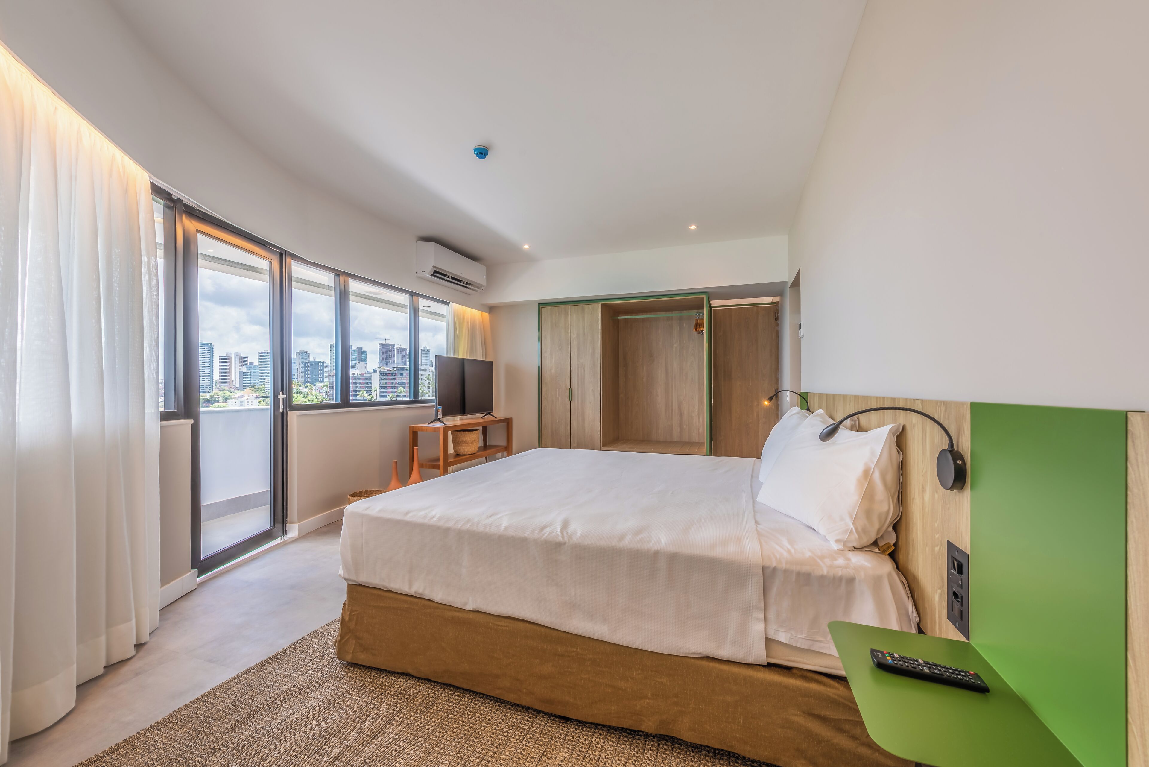 Una habitación de hotel moderna con una cama grande, televisión, escritorio verde y vista a la ciudad a través de la ventana.