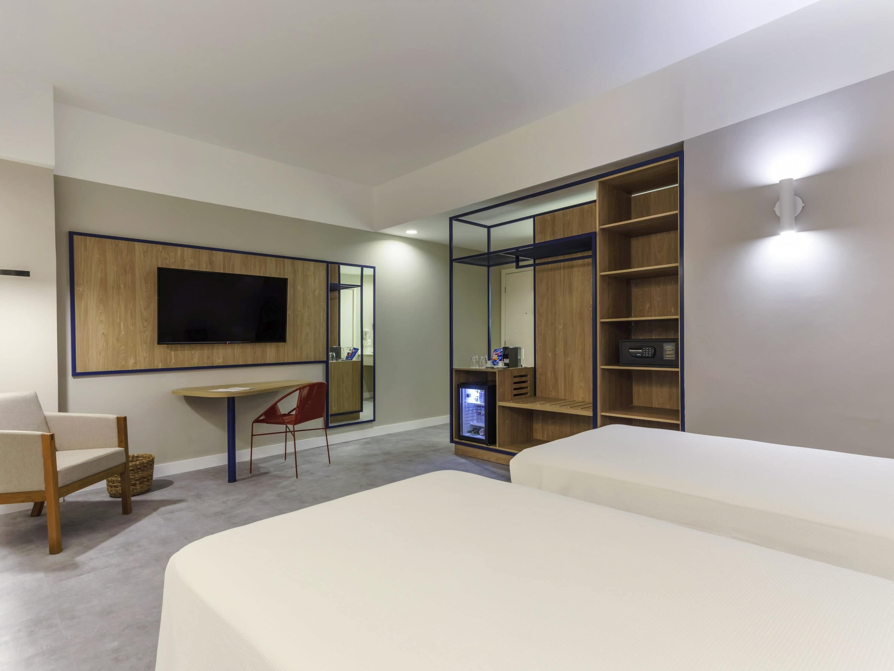 Una habitación de hotel moderna con una cama grande, un escritorio con una silla, un televisor montado en un panel de madera, un armario con estantes y un refrigerador pequeño.