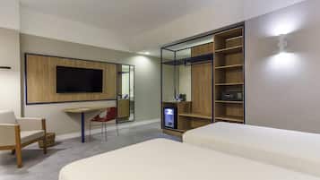Una habitación de hotel moderna con una cama grande, un escritorio con una silla, un televisor montado en un panel de madera, un armario con estantes y un refrigerador pequeño.