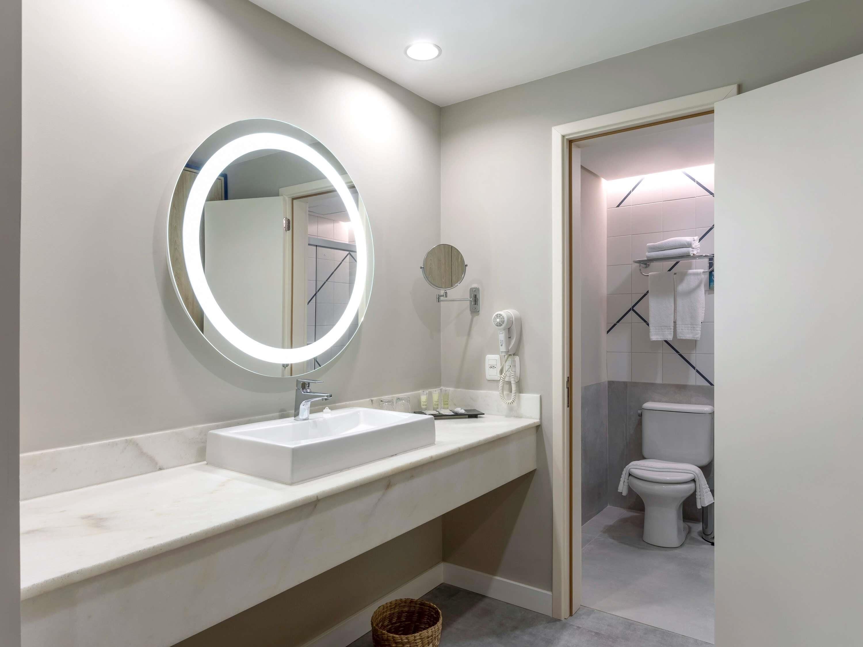 Une salle de bain moderne avec un miroir rond, un lavabo blanc, un sèche-cheveux et des toilettes visibles à travers une porte ouverte.