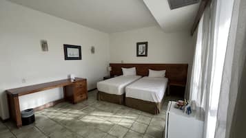 A hotel room with two beds, a desk, a trash bin, and a refrigerator.