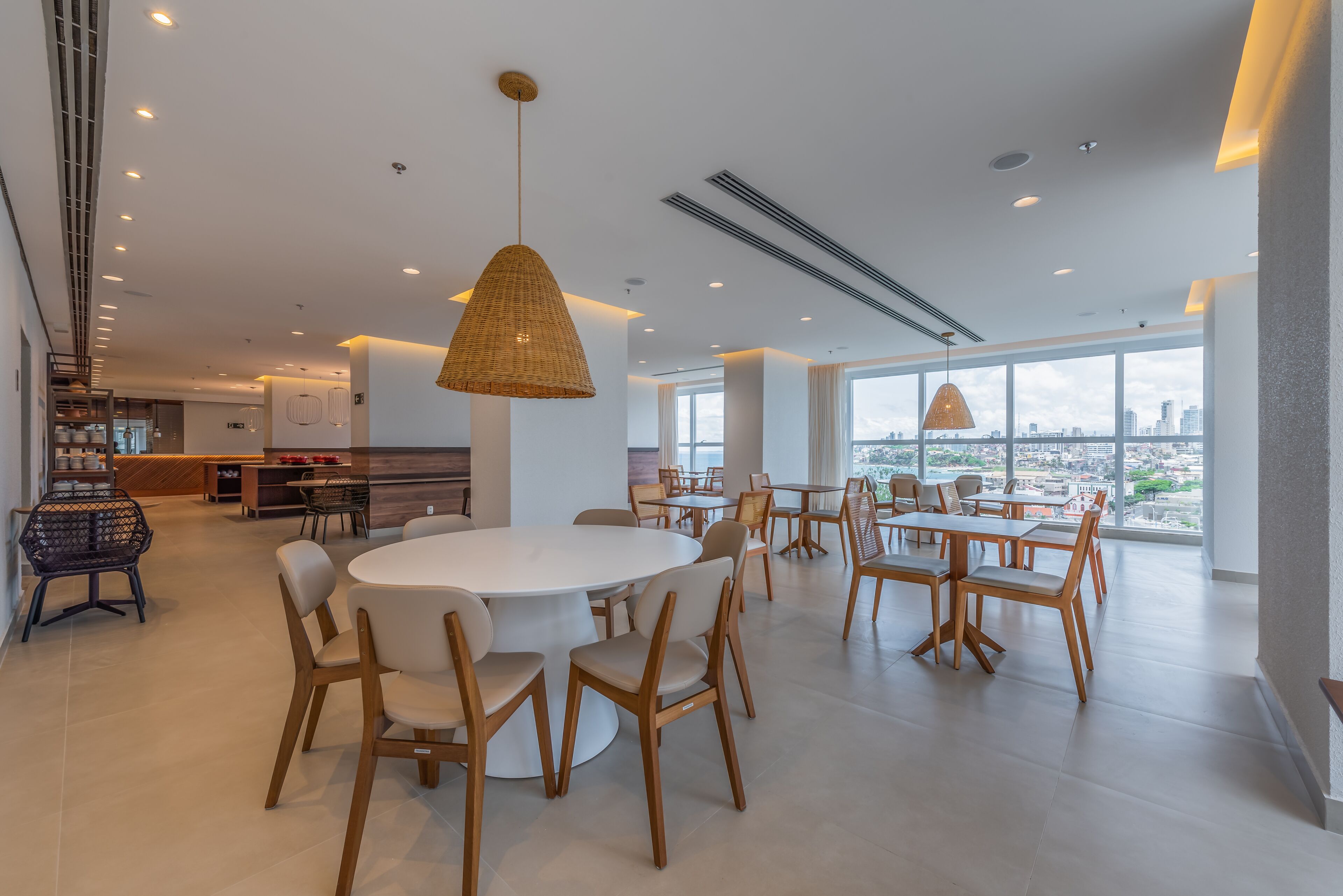 A modern dining area with round tables and chairs, large windows with a city view, and a kitchen in the background.