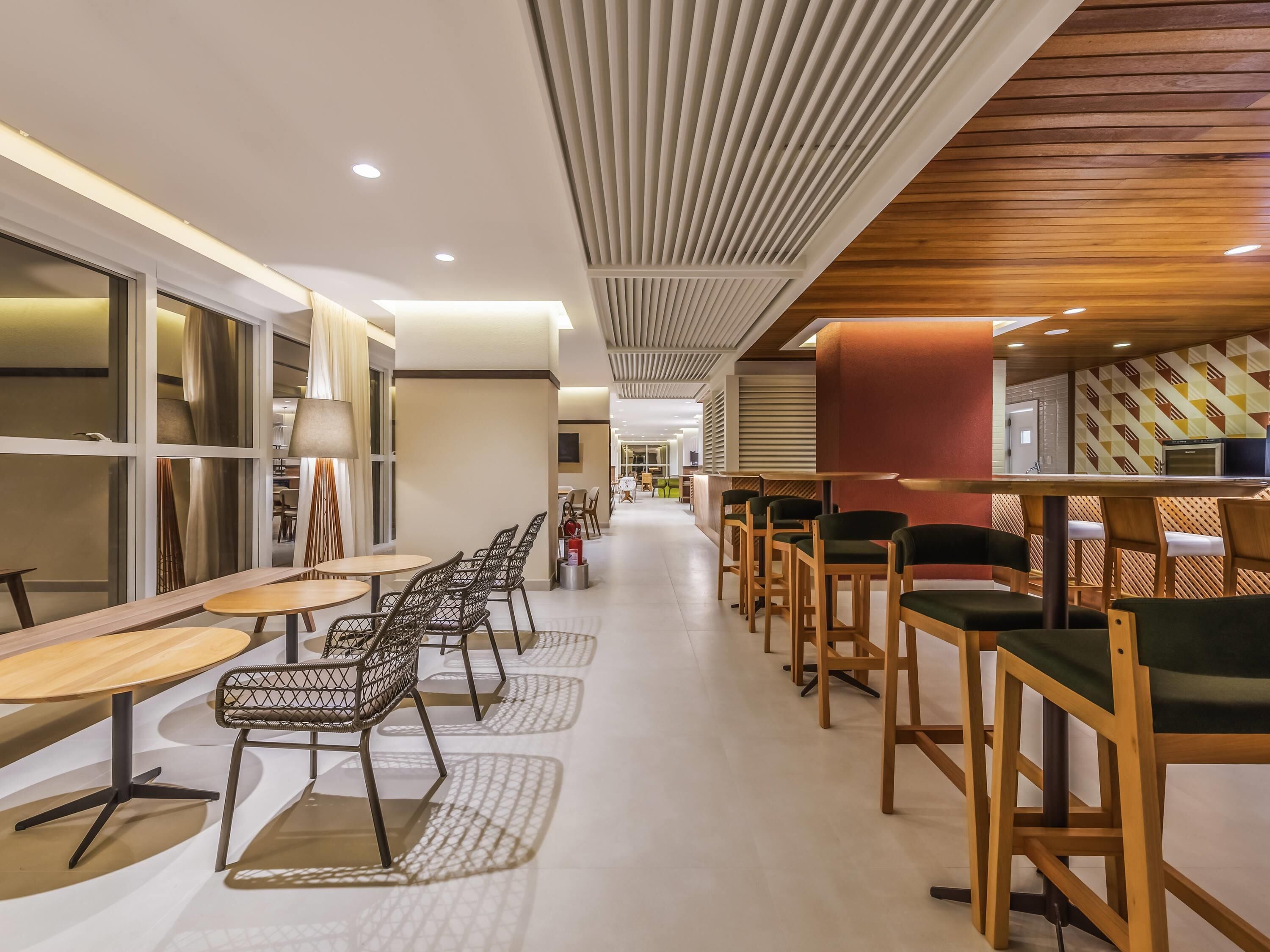 A modern interior with a long corridor lined with tables and chairs, a wooden ceiling, and a patterned wall.