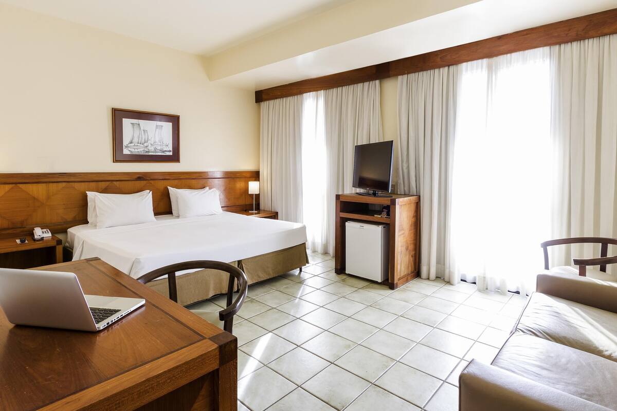 A hotel room with a bed, a desk with a laptop, a TV on a stand, and a small refrigerator.